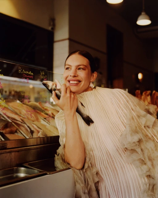 Smiling woman in ruffled dress holds a fish next to a seafood display, indoor setting, warm lighting, commercial environment.
