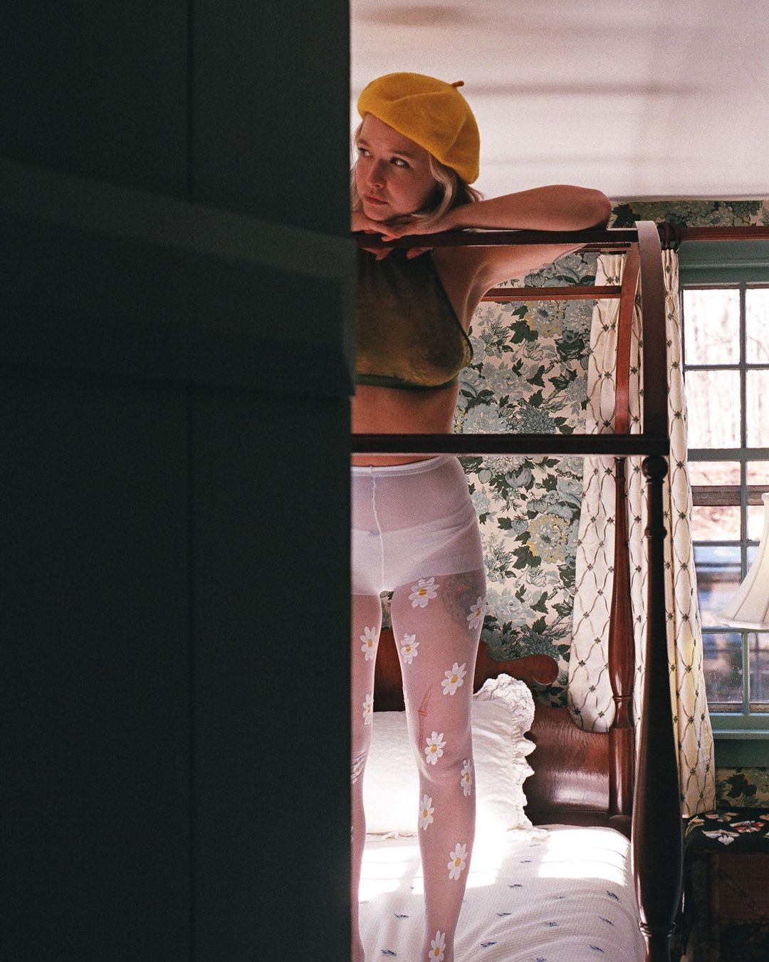 Young woman stands on a four-poster bed in a bedroom with floral wallpaper, wearing patterned tights, a halter top, and a yellow beret.
