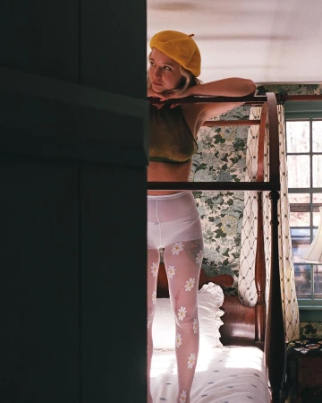 Young woman stands on a four-poster bed in a bedroom with floral wallpaper, wearing patterned tights, a halter top, and a yellow beret.