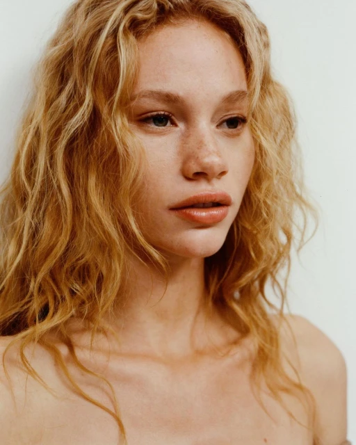 Close-up portrait of a young adult woman with blonde curly hair and freckles against a plain white background in a studio setting.