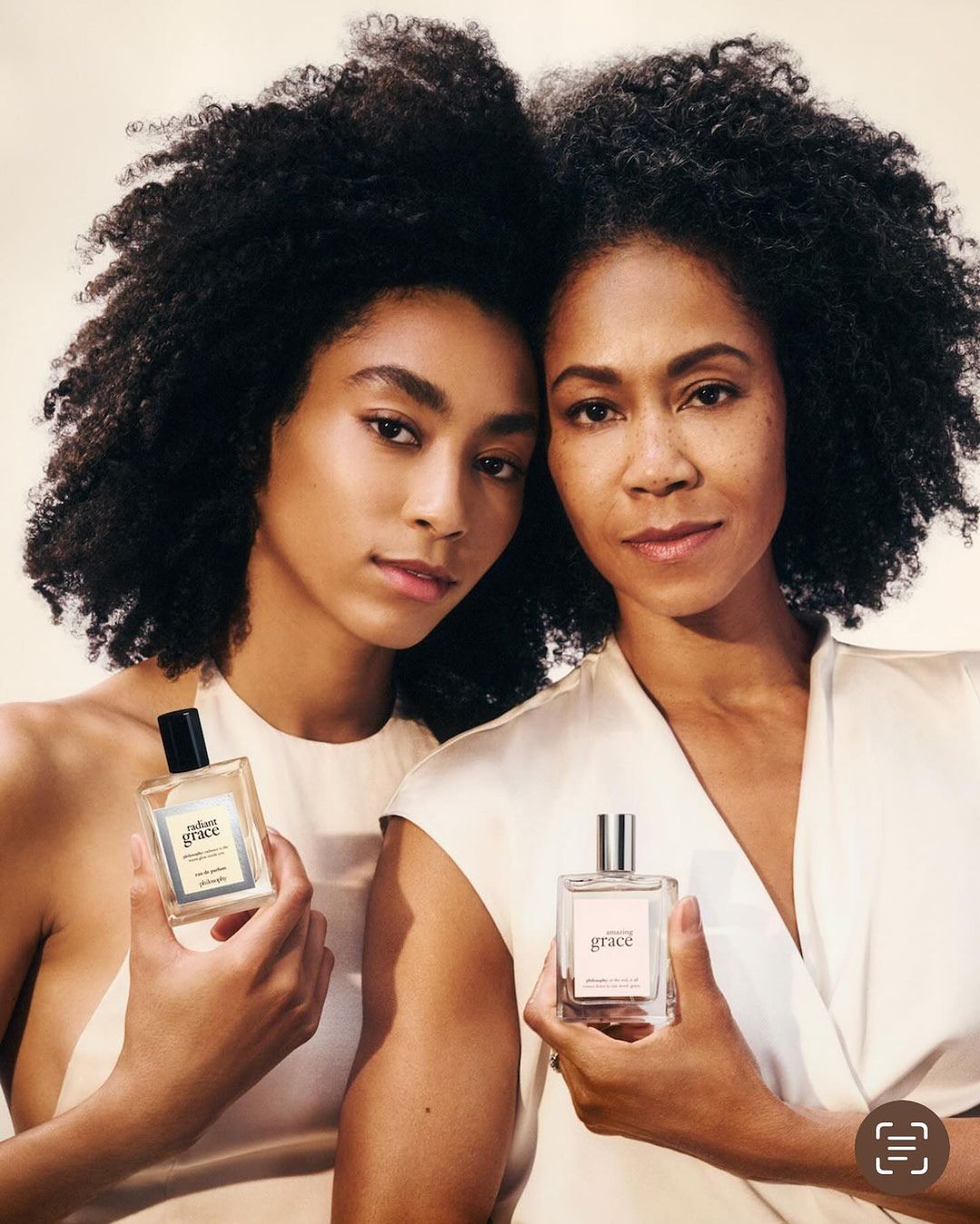 Two Black women pose in a studio setting with a bottle of perfume. They both have dark curly hair and are wearing off-white clothing.