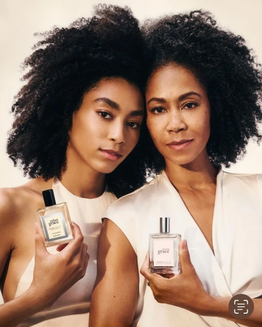 Two Black women pose in a studio setting with a bottle of perfume. They both have dark curly hair and are wearing off-white clothing.