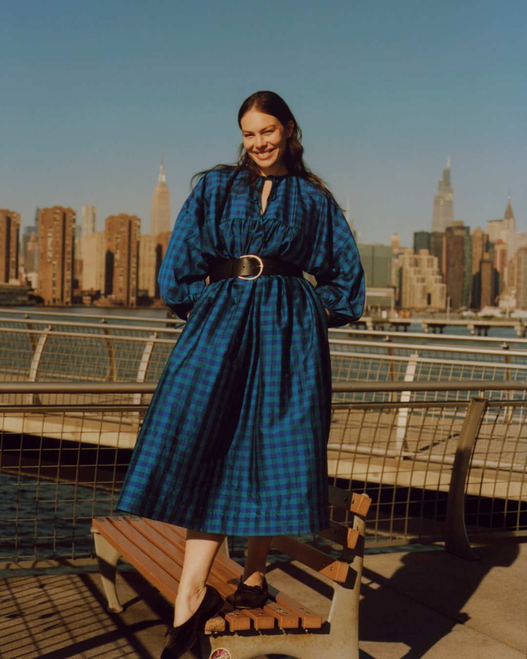 Smiling woman in a blue plaid dress with a black belt posing on a wooden park bench against a city skyline