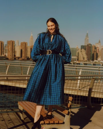Smiling woman in a blue plaid dress with a black belt posing on a wooden park bench against a city skyline