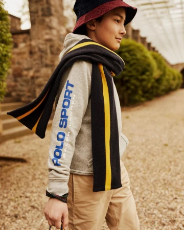 A young boy wearing a gray Ralph Lauren hoodie and a navy and yellow scarf poses outdoors on a sunny day in a natural setting.