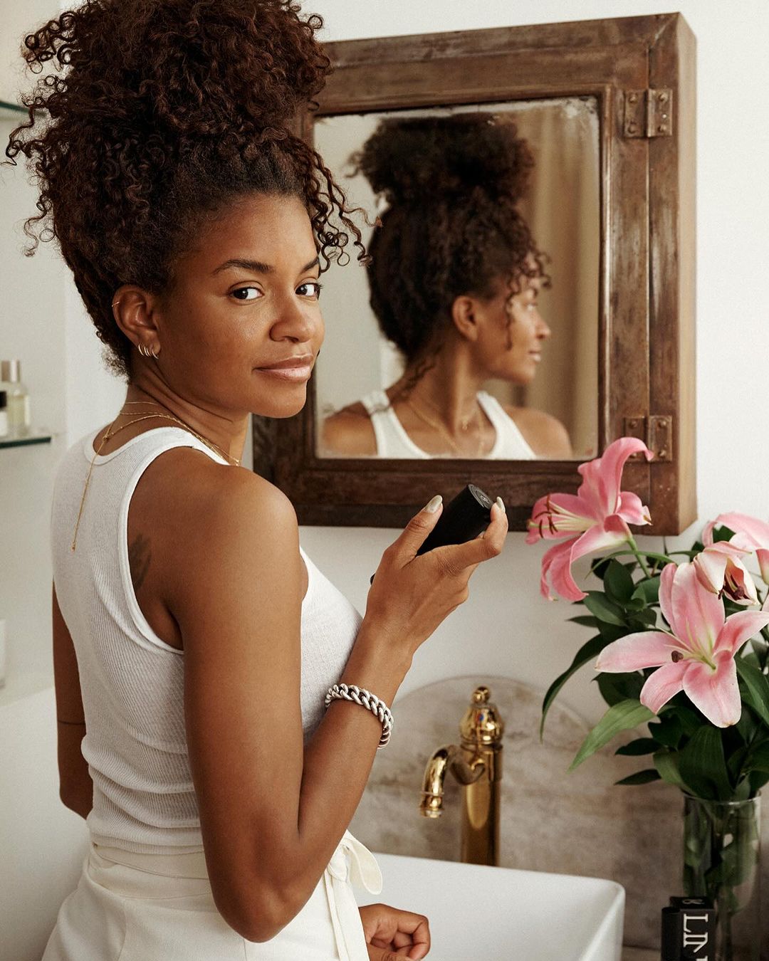 Woman with dark curly hair poses with skincare product in a bathroom, mirror reflecting her profile, accessorized with bracelet and jewelry.