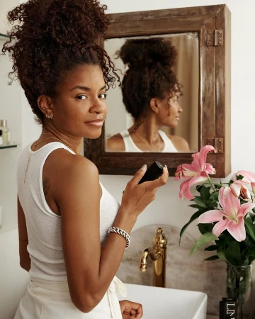 Woman with dark curly hair poses with skincare product in a bathroom, mirror reflecting her profile, accessorized with bracelet and jewelry.