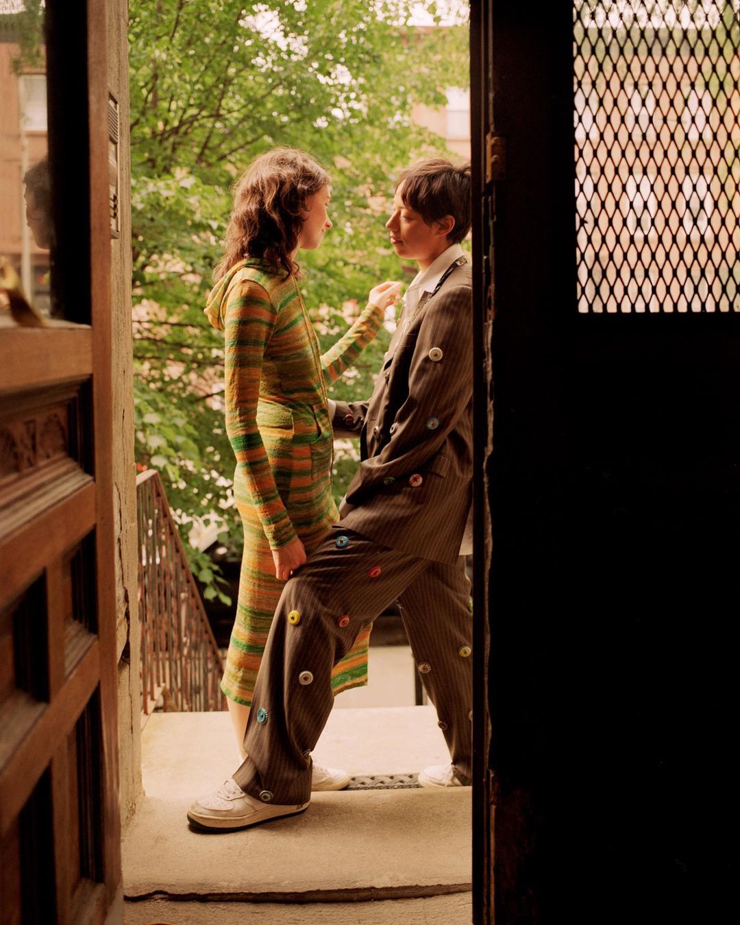 Two people stand on a stoop; one in a striped knit dress, the other in a button-covered suit with leafy trees and buildings in the background.