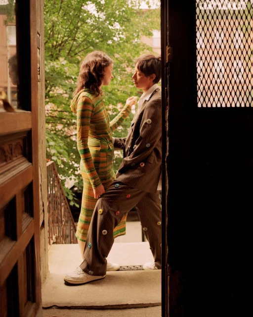 Two people stand on a stoop; one in a striped knit dress, the other in a button-covered suit with leafy trees and buildings in the background.