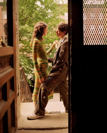 Two people stand on a stoop; one in a striped knit dress, the other in a button-covered suit with leafy trees and buildings in the background.