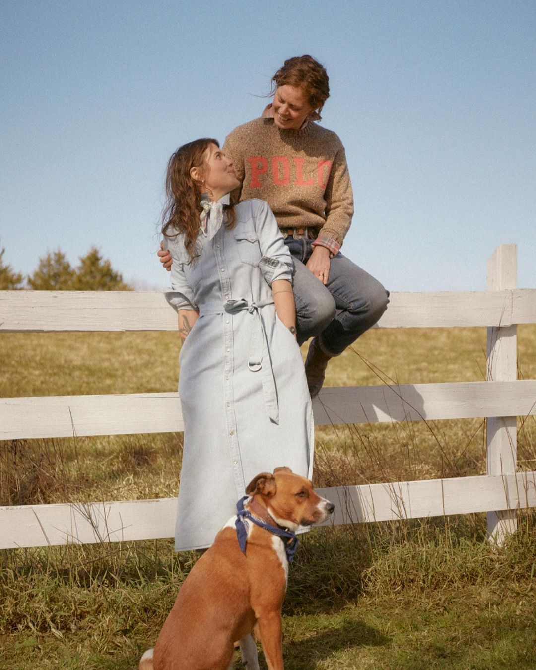 Two women in a rural setting with a dog. One woman sits on a white fence while the other stands nearby in a denim dress.