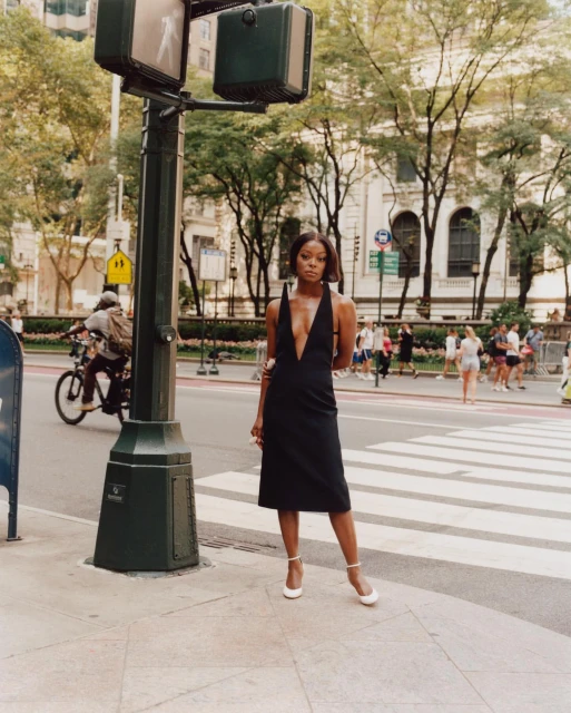 A young woman in a black dress and white heels stands on a crosswalk in an urban setting with a traffic light and city buildings.
