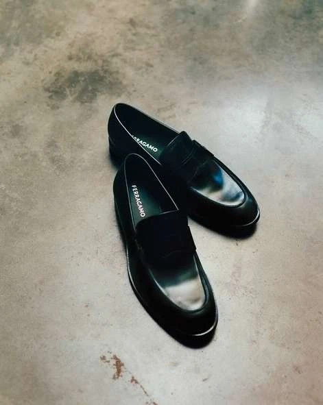 Two black leather Ferragamo loafers are displayed on a concrete surface, illuminated by neutral studio lighting, showcasing fashion footwear.