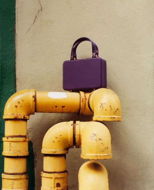 Purple handbag on yellow industrial pipes against a textured neutral wall with green trim, fashion-forward urban aesthetic.
