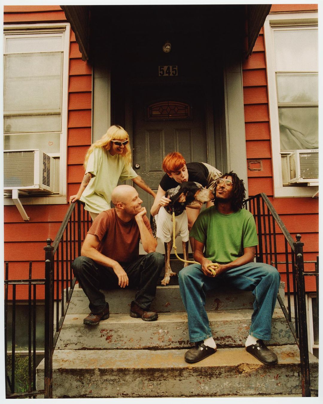 Group of five people and a dog pose on the steps of a red house with an air conditioning unit in the window and a door numbered 545.