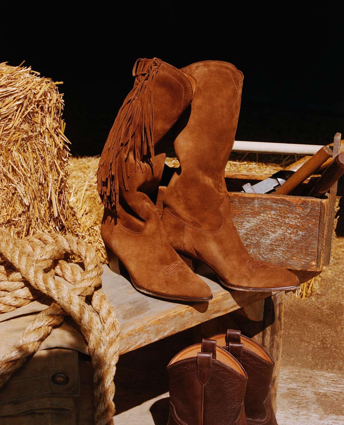 Pair of brown suede cowboy boots displayed on a wooden surface with a hay bale in the background, rustic fashion advertisement.