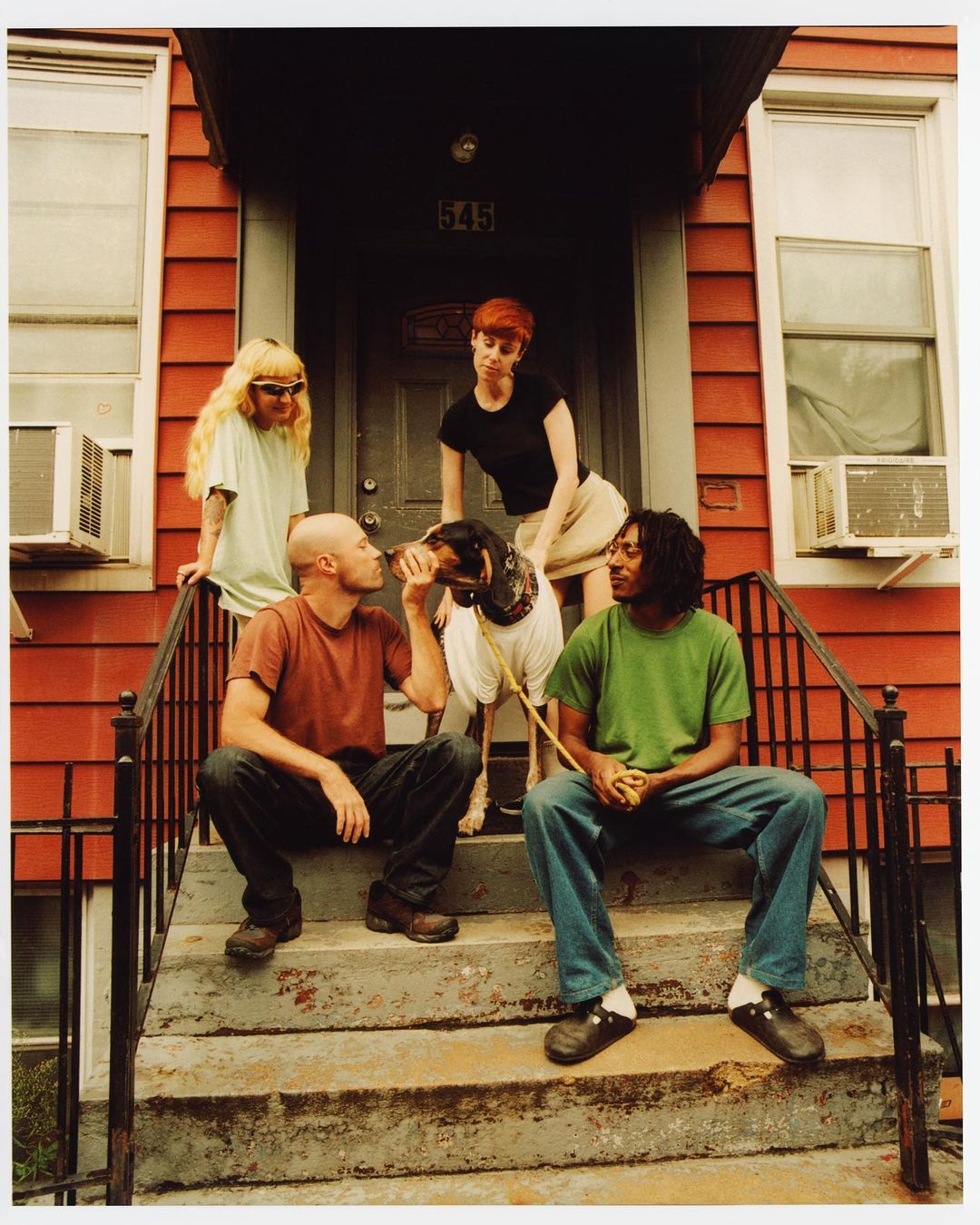 Outdoor shot of four people and a dog sitting on the steps of a red house. A man is kissing the dog, and others are watching.
