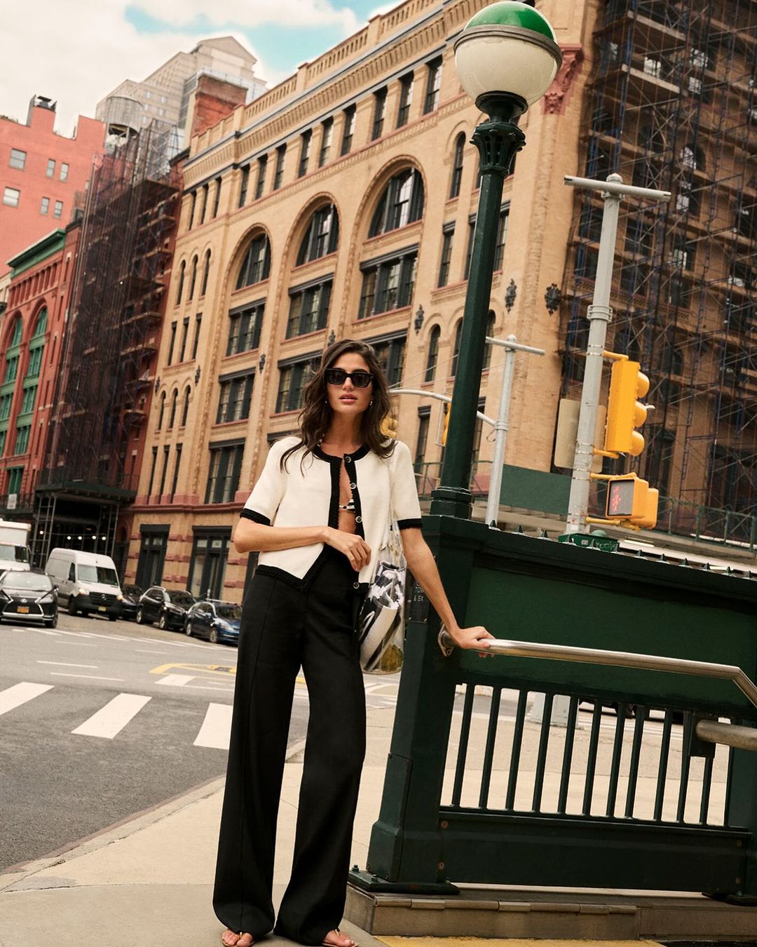 A brunette woman poses in a cream jacket and black pants near a subway entrance in an urban setting, with buildings in the background.