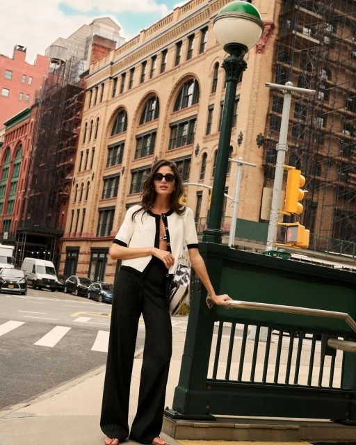 A brunette woman poses in a cream jacket and black pants near a subway entrance in an urban setting, with buildings in the background.