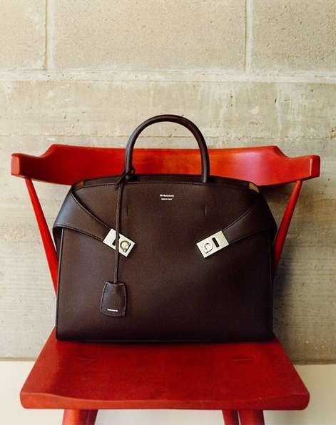 Brown leather handbag with silver hardware on a red wooden chair against a neutral wall, showcasing fashion accessory.