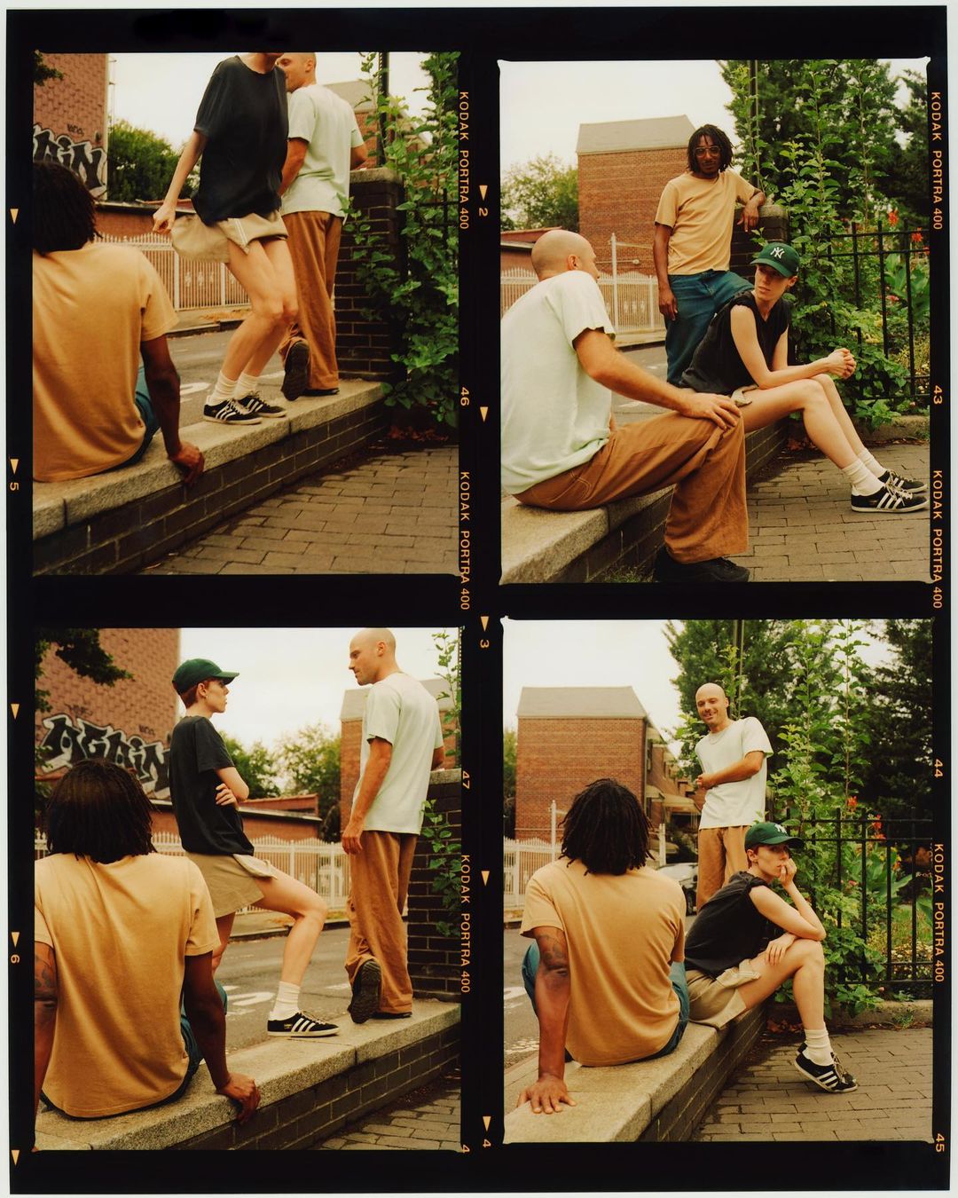 Group of young adults are hanging out on a city street with brick buildings, wearing casual clothes, sitting on a ledge, and posing.