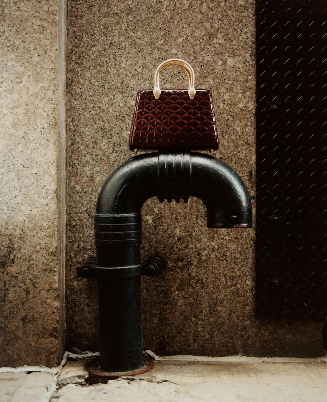 Burgundy purse with gold handle sits atop a black pipe against a textured beige wall with black metal panel, close-up.