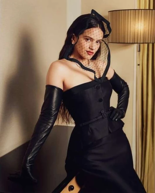 Rosalia poses in a black strapless dress with long leather gloves and a netted veil, set against a neutral wall with a lamp.