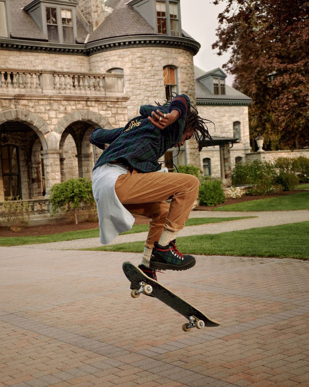 Young adult skateboarding in front of a stone building. The skateboarder is mid-air, wearing casual clothing on a brick surface outdoors.