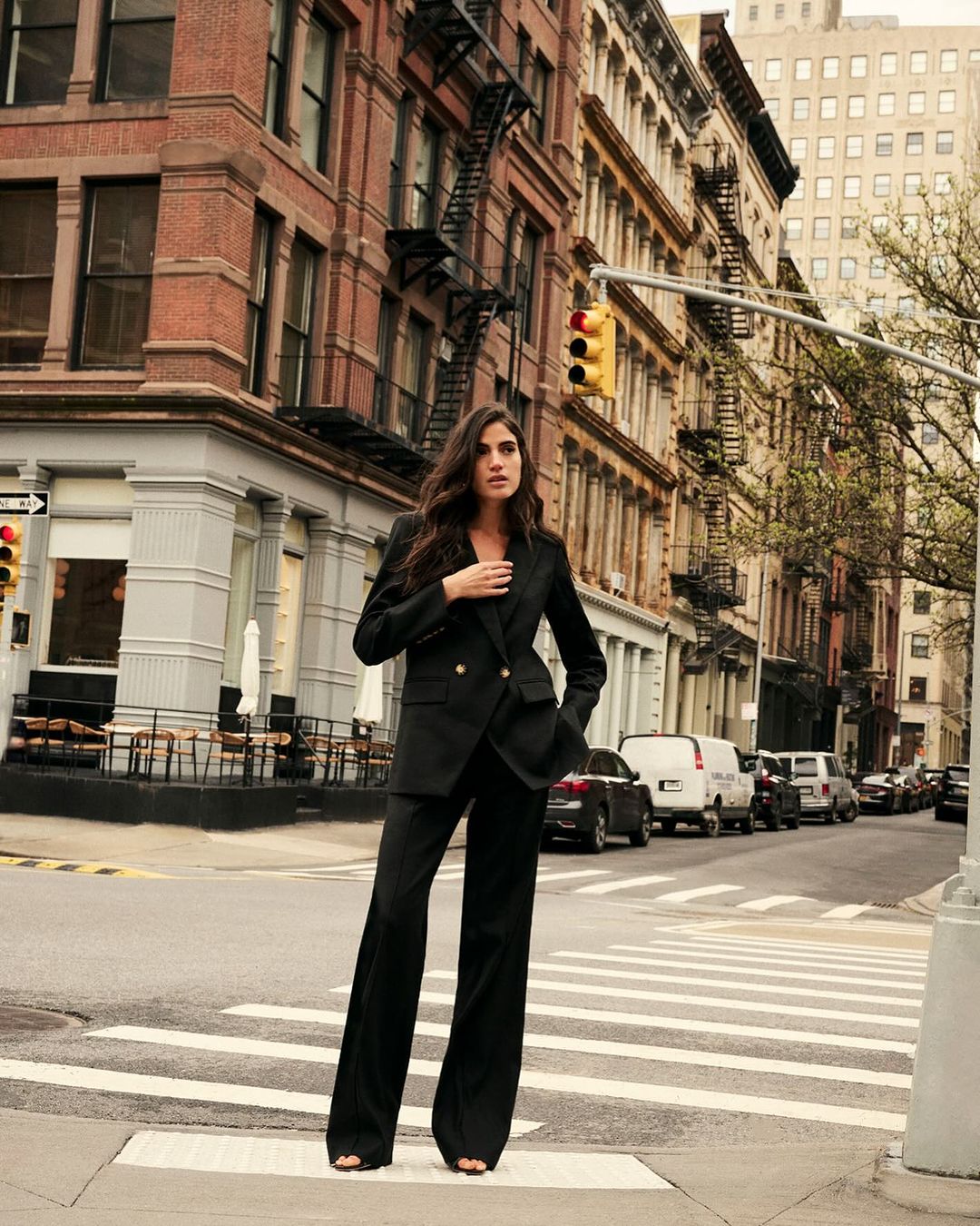 Woman in a black pantsuit walks across a crosswalk in an urban setting with brick buildings and cars in the background.