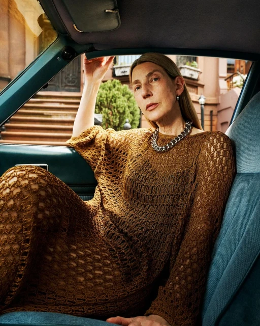 Adult woman in an open-knit brown dress sits in the driver's seat of a blue car in front of a brick apartment building.