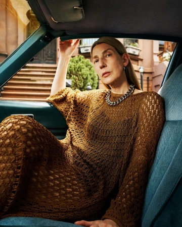 Adult woman in an open-knit brown dress sits in the driver's seat of a blue car in front of a brick apartment building.