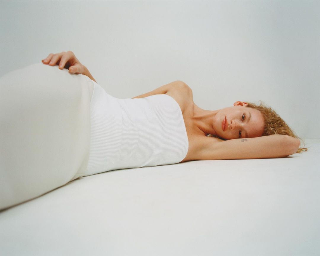Woman with curly red hair lying on her side wearing a white top and white skirt in a studio with a white background.