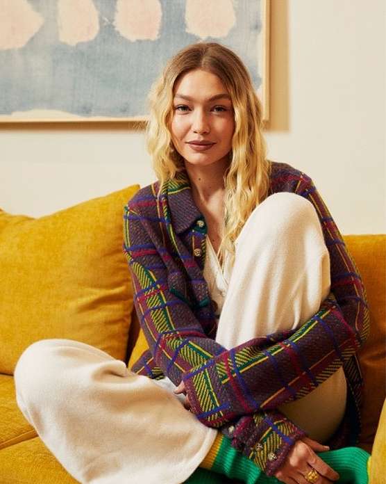 Blonde woman in a plaid jacket sits on a yellow sofa with a painting visible in the background, posing in a studio environment.