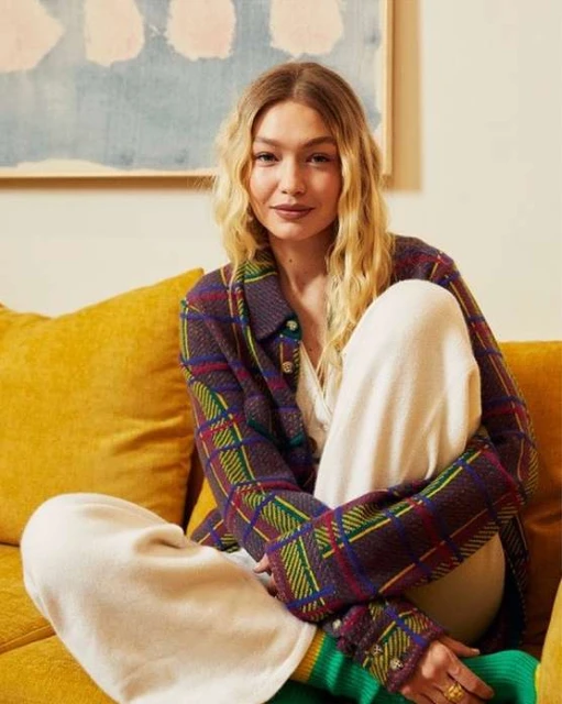Blonde woman in a plaid jacket sits on a yellow sofa with a painting visible in the background, posing in a studio environment.