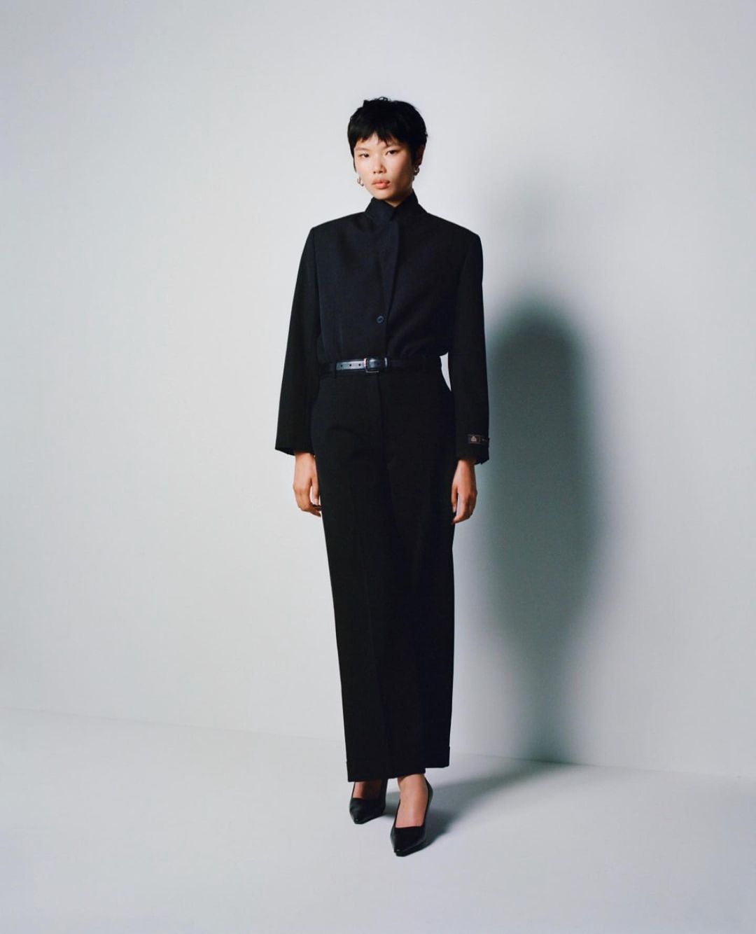 A young woman stands in a photo studio wearing a black blouse, wide-leg pants, a thin belt, and high heels against a white backdrop.