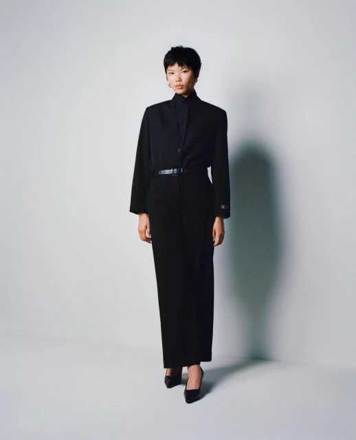 A young woman stands in a photo studio wearing a black blouse, wide-leg pants, a thin belt, and high heels against a white backdrop.