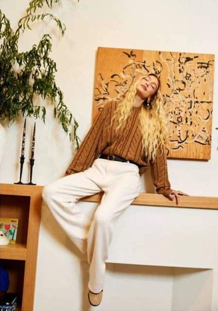 A blonde woman poses indoors in a brown knit sweater and white pants, seated on a shelf, next to plants and a painting.