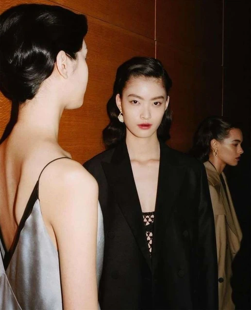Three women in a dimly lit setting. One woman faces forward in a black blazer, another is seen from behind in a light-colored dress.