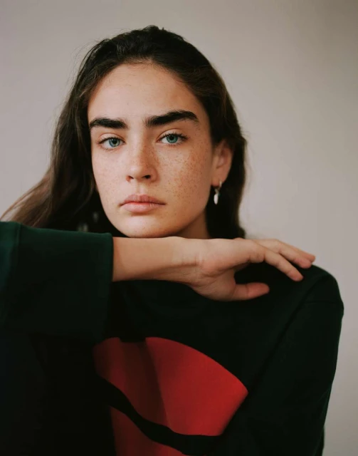 Portrait of a young woman with freckles, dark hair, and a serious expression wearing a black sweater with a red graphic. Neutral studio backdrop.