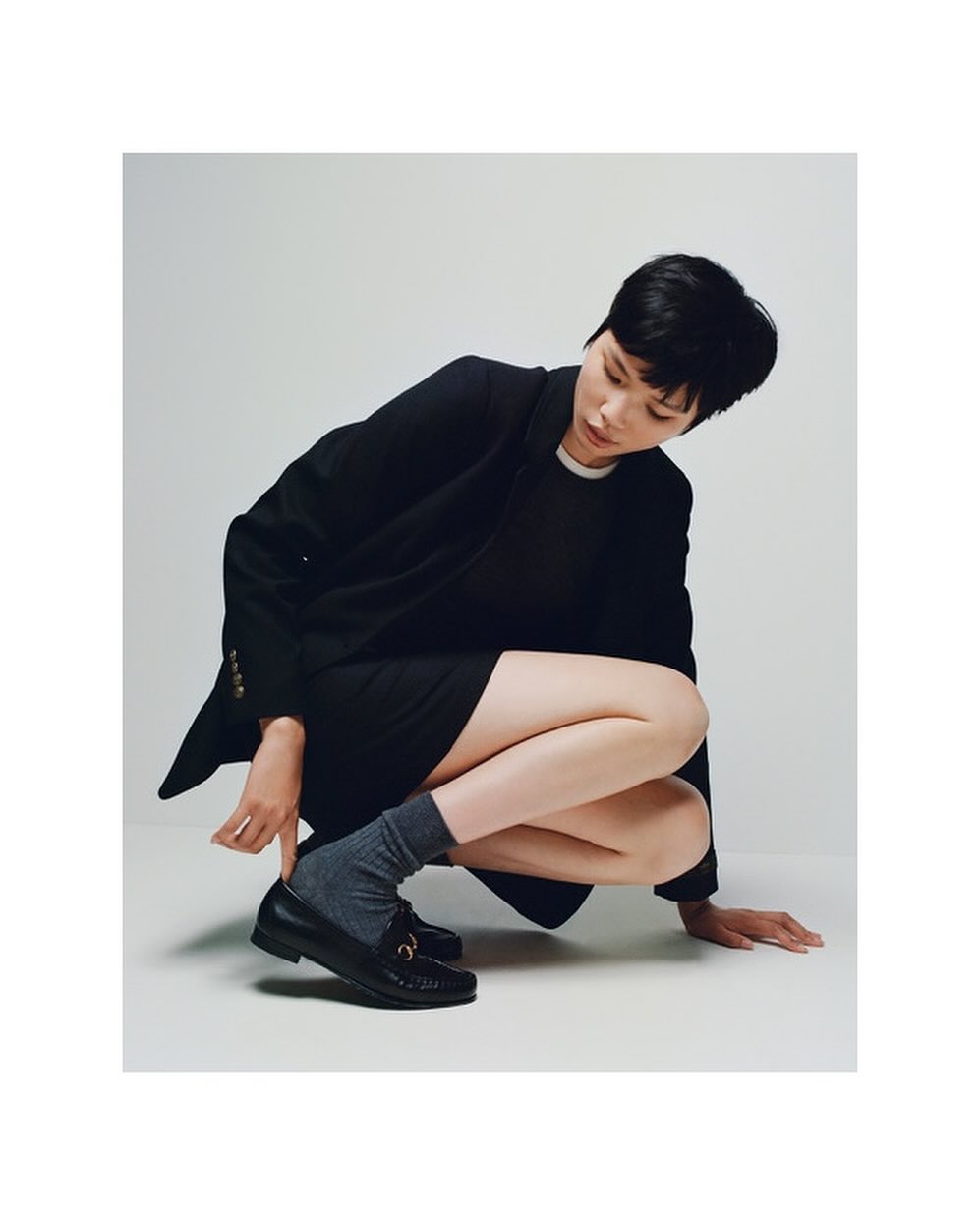 Woman in a dark blazer and skirt, crouching with one hand adjusting her black loafer with socks, in front of a white background.