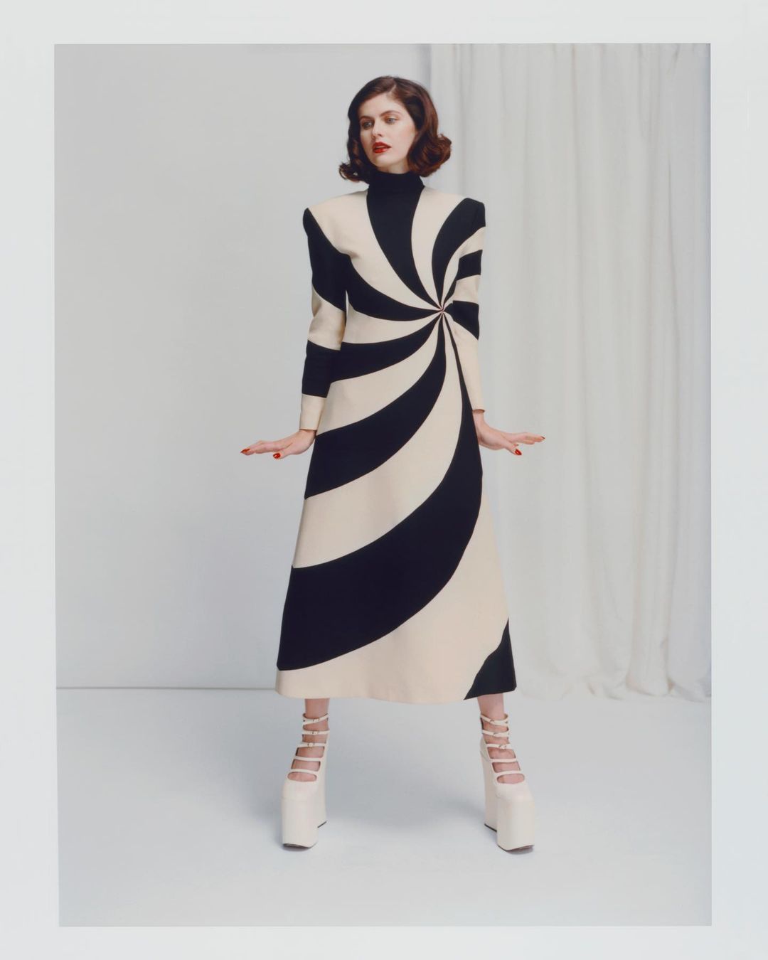Fashion photo of a woman wearing a black-and-white spiral-patterned dress and white platform shoes in a photo studio setting.