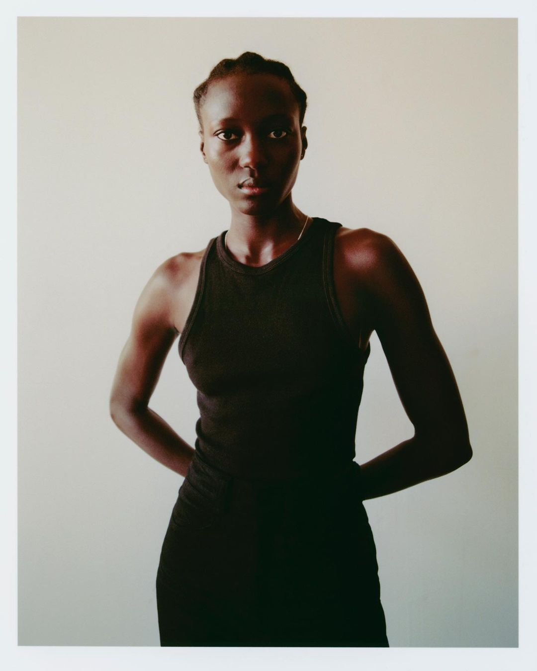 Portrait of a young Black woman in a black tank dress against a neutral background, hands clasped behind her back.
