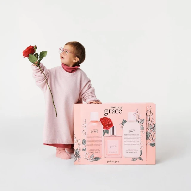 A young child in a pink outfit holds a red rose next to a pink gift box set of 'amazing grace' fragrance products.