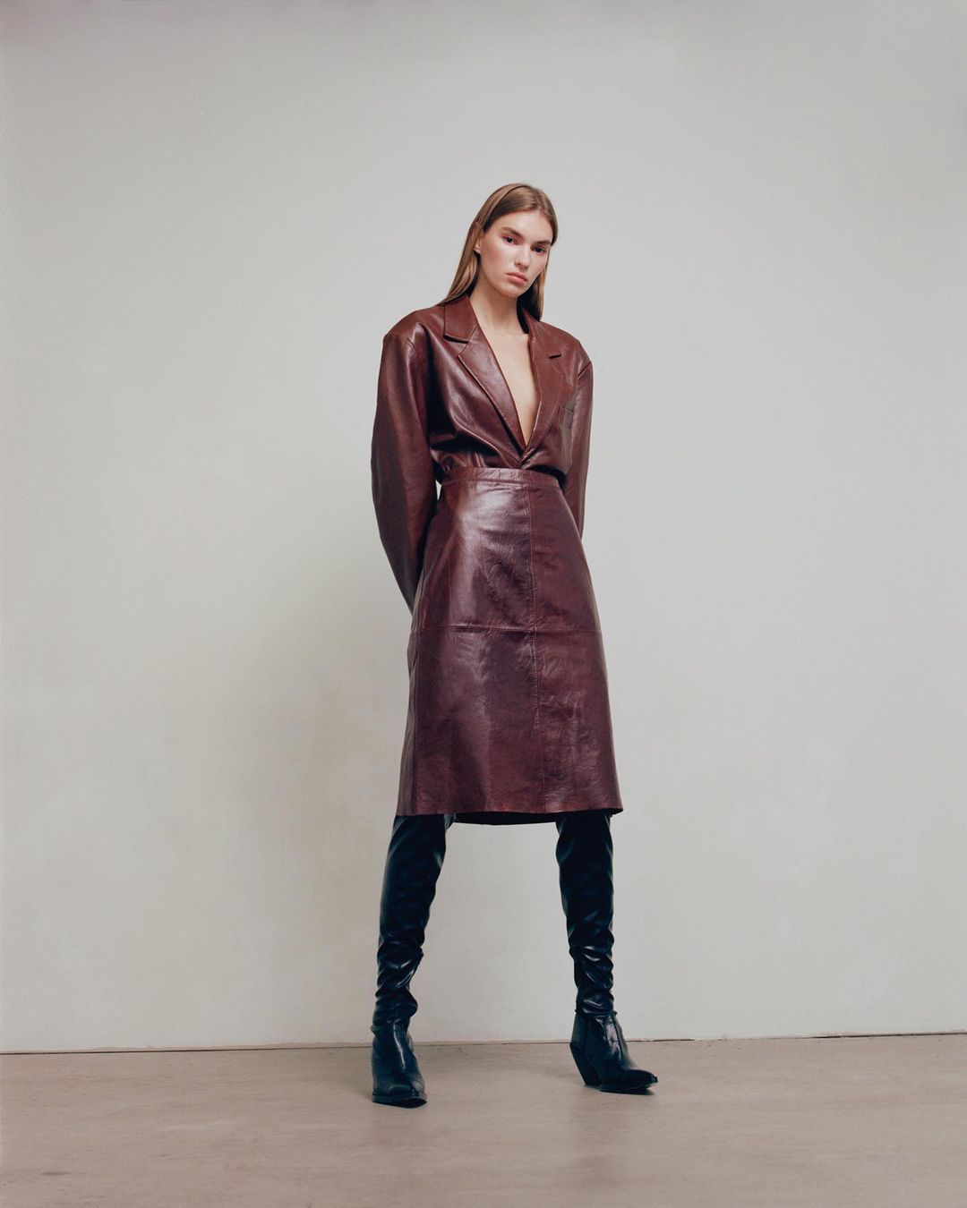 Fashion shot of a woman in a brown leather dress and black boots standing against a neutral background in a studio setting.