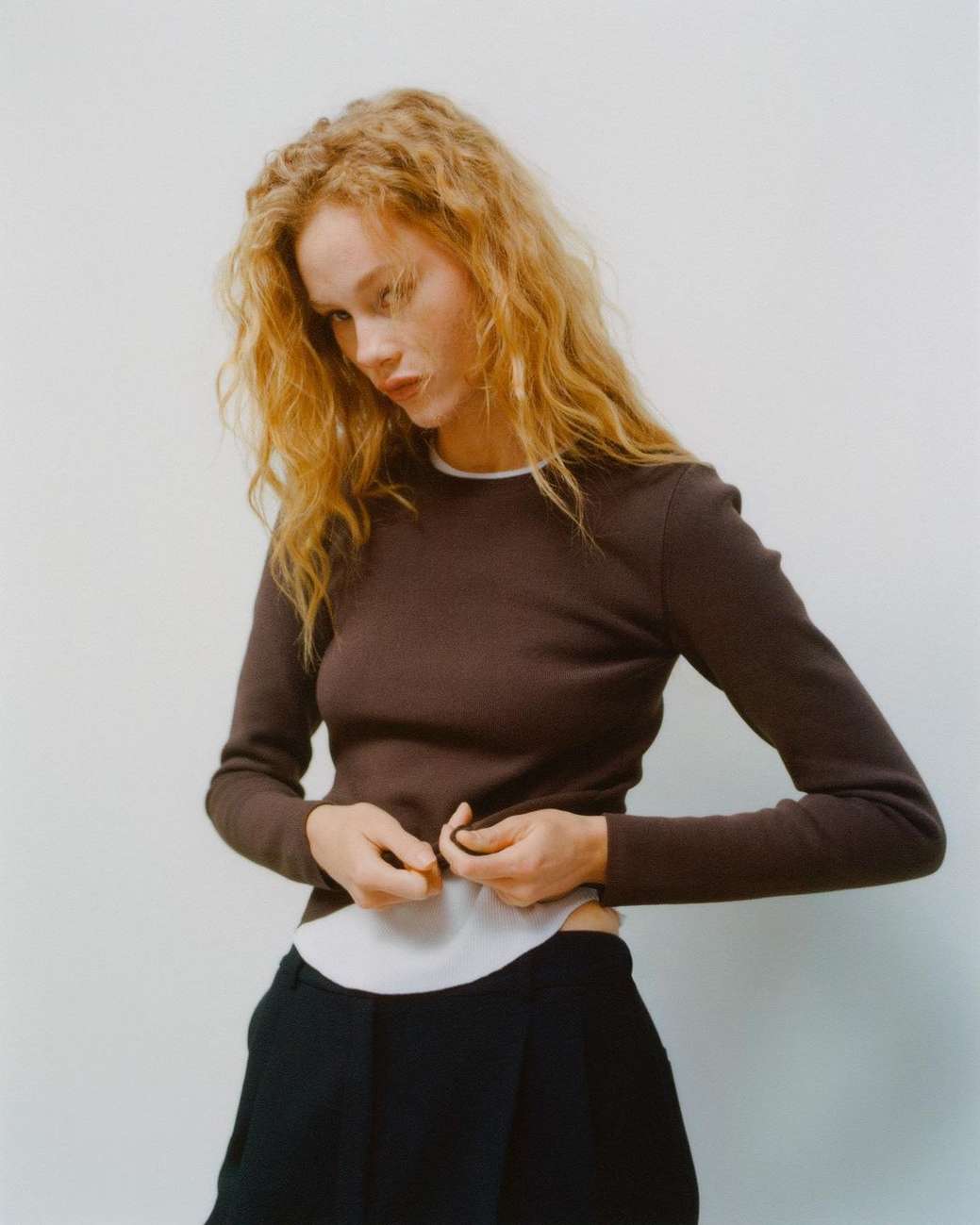 Woman in a brown long-sleeve shirt adjusts her white undershirt. She has light skin and curly hair, set against a light background.