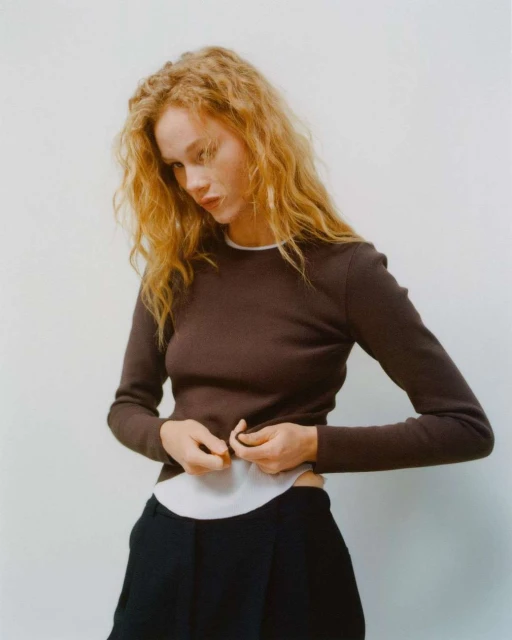 Woman in a brown long-sleeve shirt adjusts her white undershirt. She has light skin and curly hair, set against a light background.