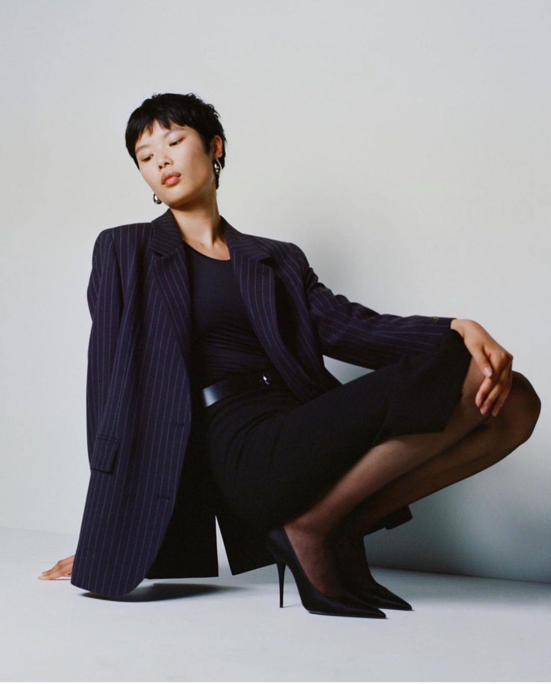 Woman in pinstripe blazer and black heels squats in front of a white backdrop in a fashion photography shot. She wears simple silver earrings.