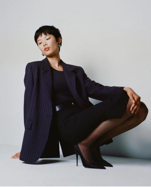 Woman in pinstripe blazer and black heels squats in front of a white backdrop in a fashion photography shot. She wears simple silver earrings.
