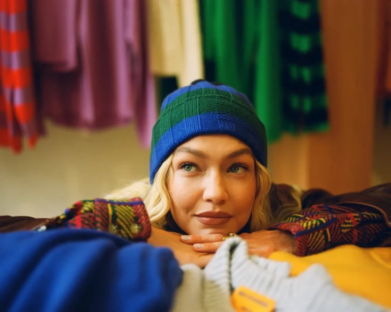 Blonde woman in a blue and green beanie leans on a pile of folded sweaters in a closet with colorful hanging garments.