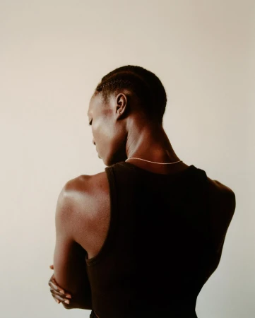 Portrait of a Black woman from behind wearing a dark sleeveless top and thin silver necklace against a plain off-white background